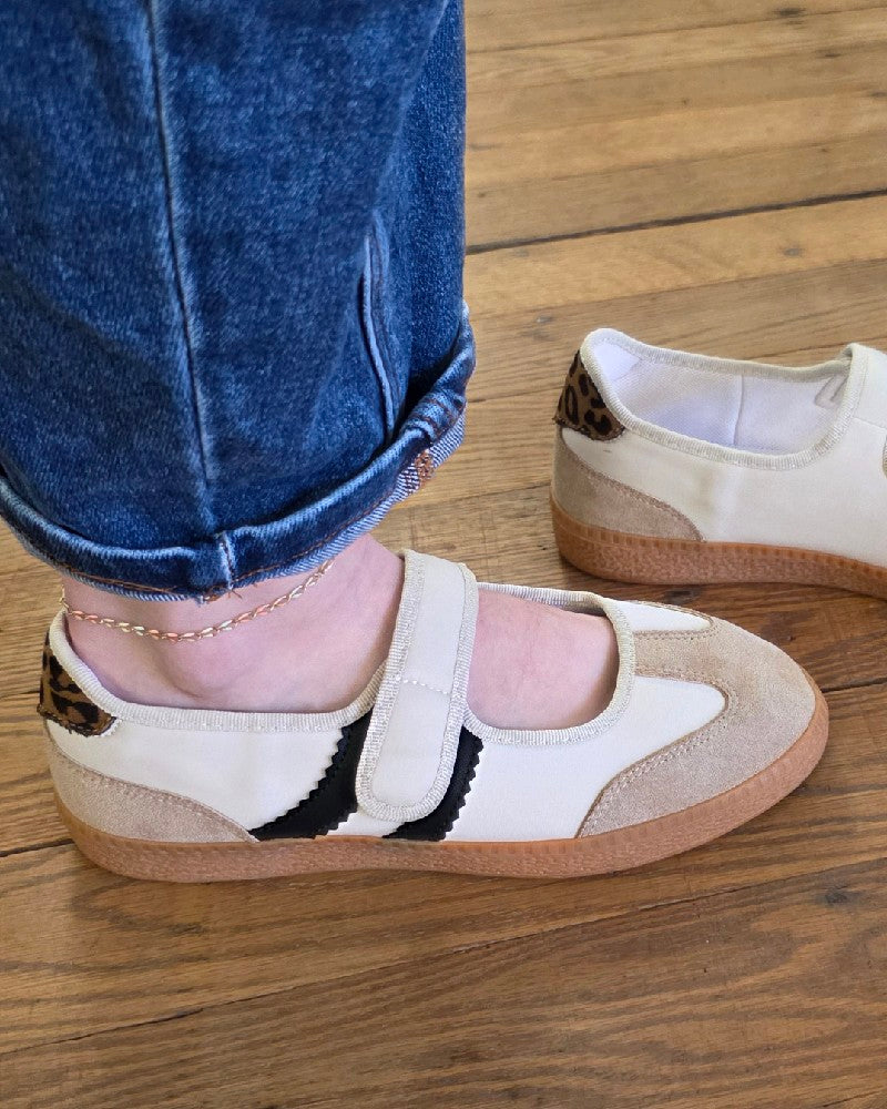 Wearing rolled-up blue jeans, a person stands on a wooden floor in the Corkys Calm Down Sand Combo Slip on Shoe—a women’s cream Mary Jane sneaker featuring a tan sole and black detail that brings a fresh twist to the classic style; another shoe can be seen in the background.