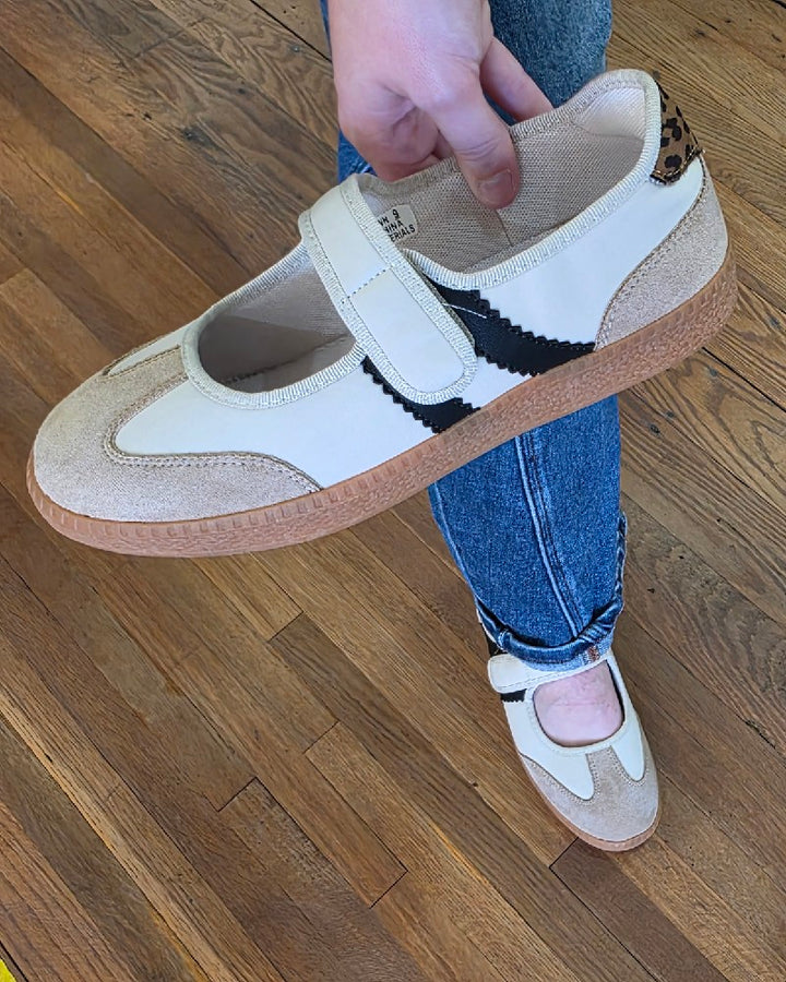 Someone wearing blue jeans is holding a Corkys Calm Down Sand Combo Slip On Shoe—this women’s slip-on features a beige upper with a gum sole and black zigzag detail; one shoe is worn while the other is held above a wooden floor.