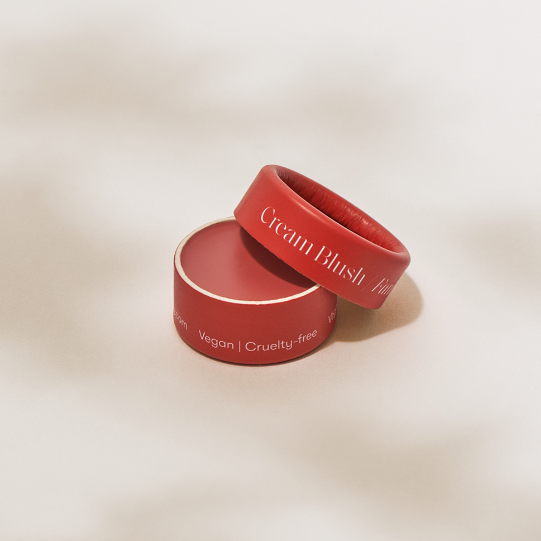 A red, circular container of Minori's Zero Waste Cream Blush Mini - Scarlet sits with its lid partially off, showing the blush inside and labeled “Vegan | Cruelty-free”—ideal for travel or eco-conscious makeup lovers.