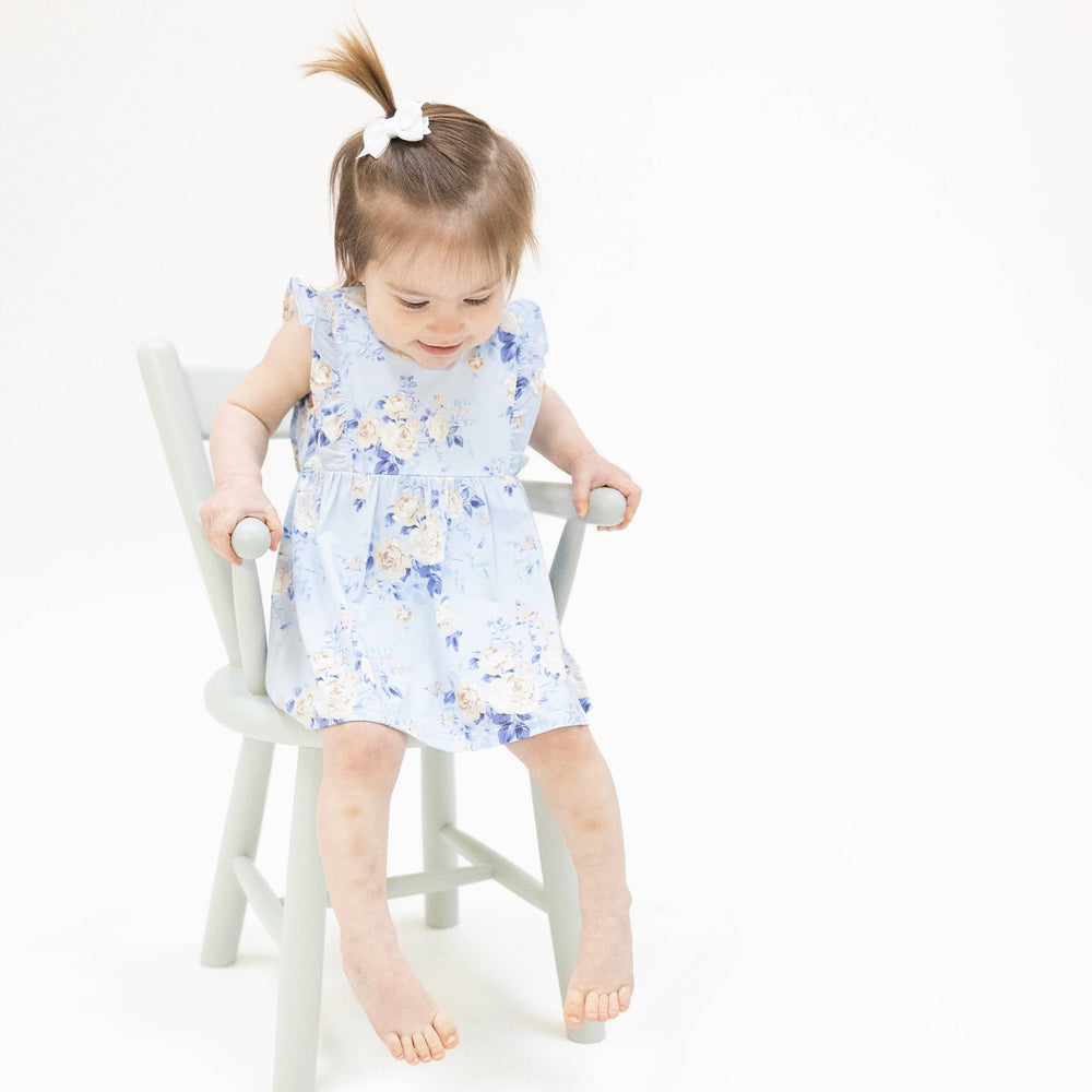 A young girl with light brown hair in a ponytail wears the Ashley Rose Floral - Ruffle Bodysuit Dress by Angel Dear, sitting on a light gray chair with her feet dangling, against a plain white background.