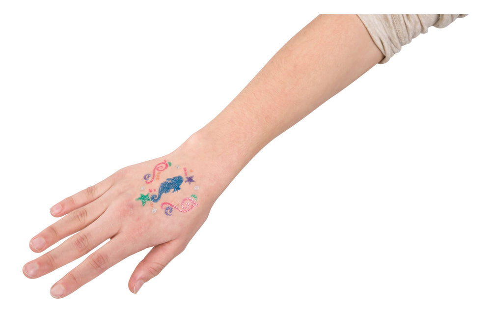 A left hand displays dazzling mermaid-themed tattoos—dolphin, stars, heart, and swirls—created with Toysmith Mermaid Tattoo Gel Pens (Set of 6). The colorful glitter designs shine against a white background.
