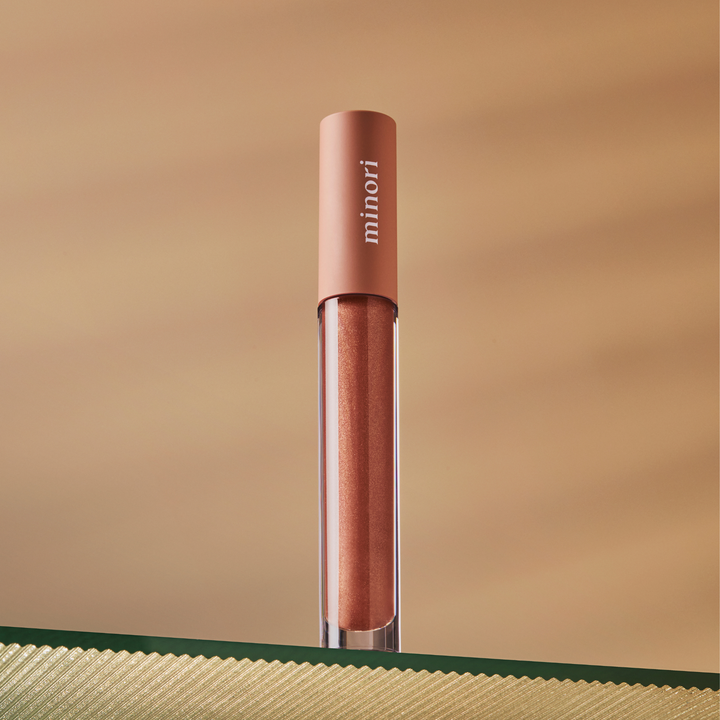 A metallic rose gold tube of Minori Lip Gloss - Cozy stands upright on a textured green and gold surface against a soft, gradient beige background.