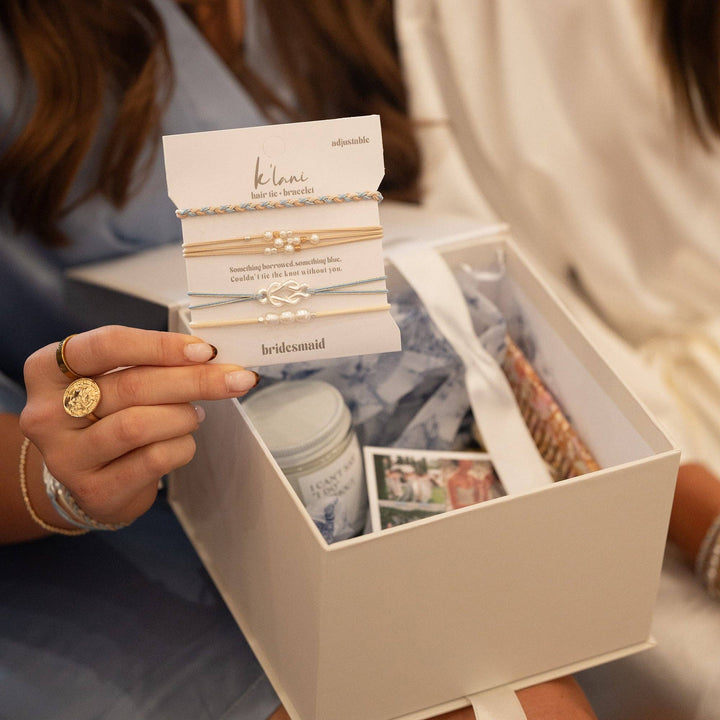 A woman holds a card featuring K’lani hair tie bracelets in the “Bridesmaid” Cream &amp; Blue with Pearl Accent design above an open white gift box filled with thoughtful items like a candle, photos, and ribbon—an elegant bridesmaid gift capturing every special detail.