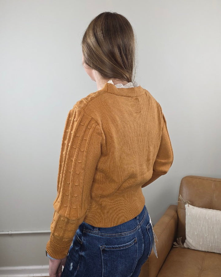 A woman with light brown hair in a ponytail wears the Heimish Elizabeth Lace Edge Cardigan and blue jeans, standing with her back to the camera in a modern living room with neutral decor.