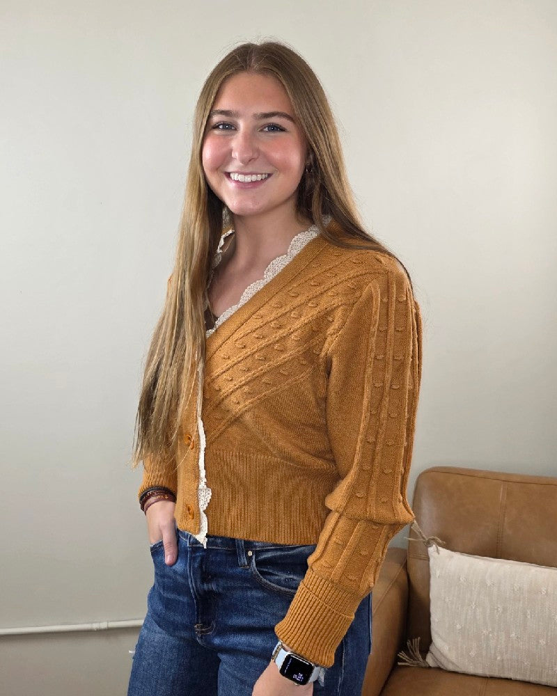 A young woman with long brown hair, wearing the Heimish “Elizabeth Lace Edge Cardigan” in mustard yellow and jeans, stands in a living room and smiles at the camera. She has one hand in her pocket and wears a smartwatch.