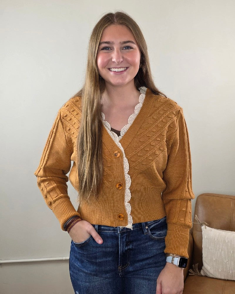 A young woman with long brown hair smiles while wearing the Heimish “Elizabeth Lace Edge Cardigan” in mustard yellow, styled with blue jeans. She stands indoors in front of a beige wall and a brown chair.