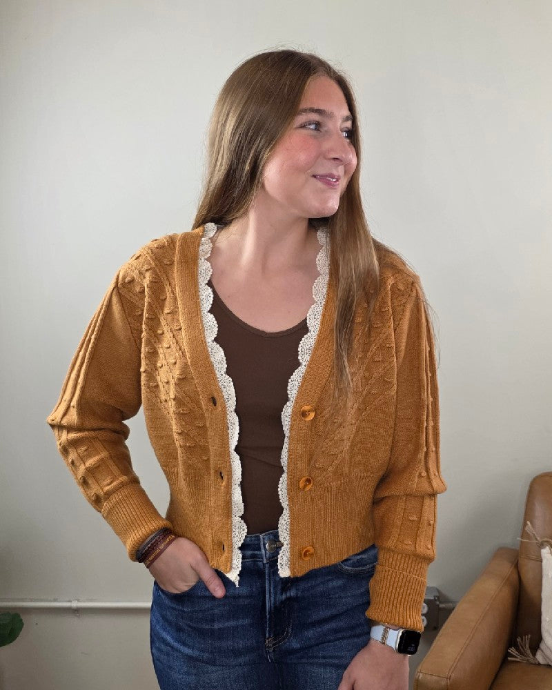 A woman with long brown hair stands indoors, smiling softly as she looks to the side, wearing the Heimish Elizabeth Lace Edge Cardigan over a brown top and blue jeans.