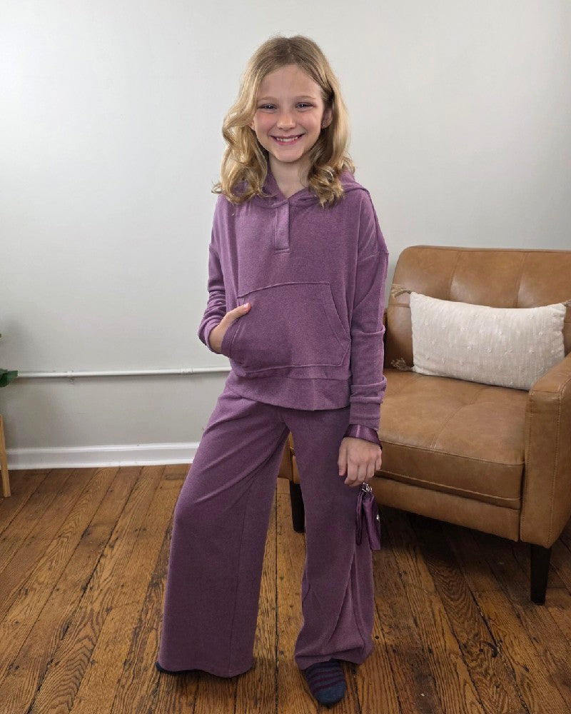A girl wearing a purple 90 Degree by Reflex Ellen Tween Girls Snap Hoodie, Wide Leg Pants and Wristlet Set, holding a small coordinated wristlet.