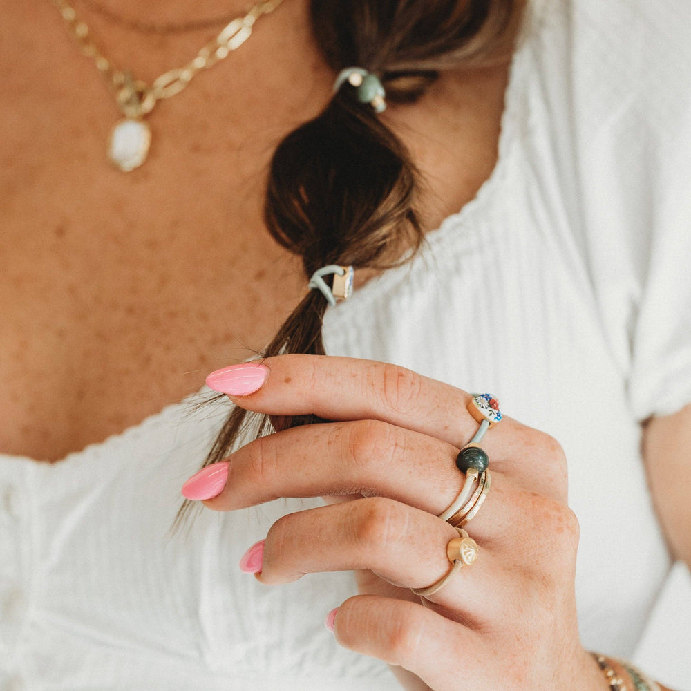 A woman with pink nails holds her braided hair, which is adorned with K’lani hair tie bracelets in the “Terra” Earth Tones Hair Tie Rings. She complements her look with gold stretchy rings, a gold necklace, and a buttoned white blouse.