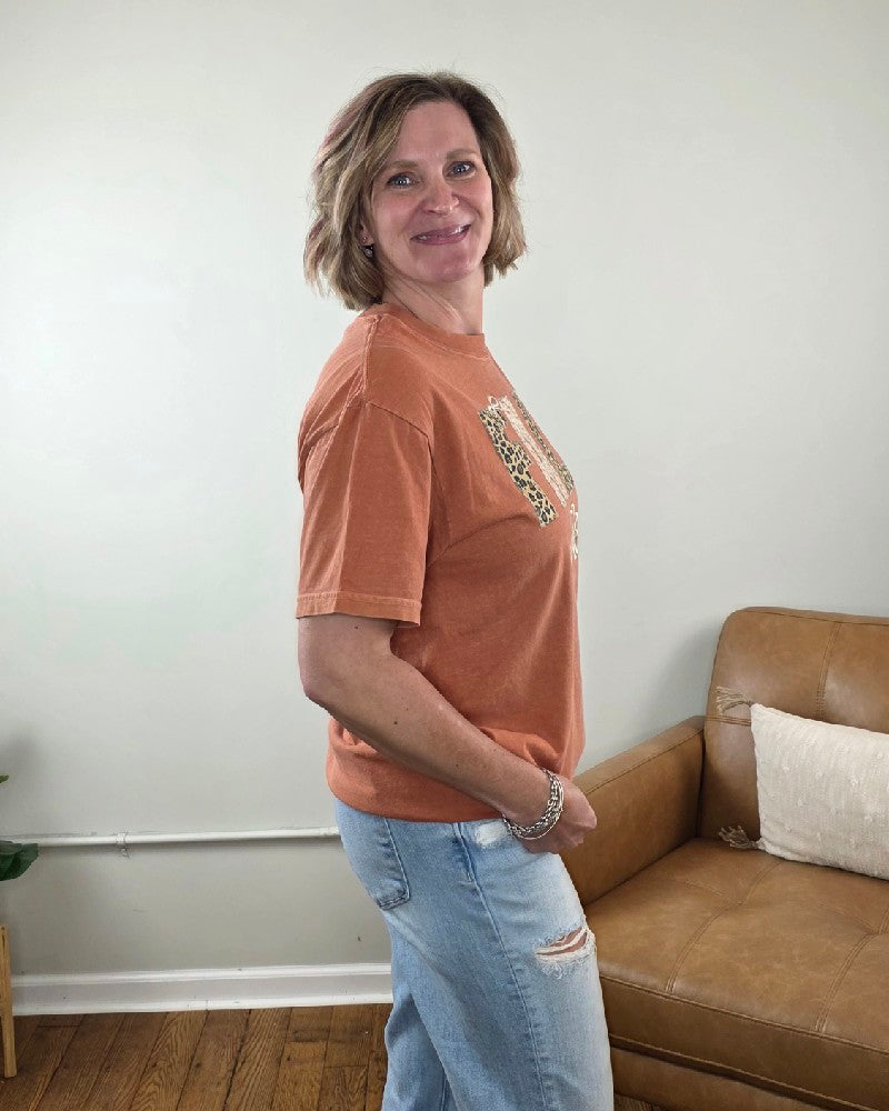 A woman wearing a rust-colored Fall Vibes Applique and Embroidered Tee with a leopard-print design and light-wash distressed jeans.
