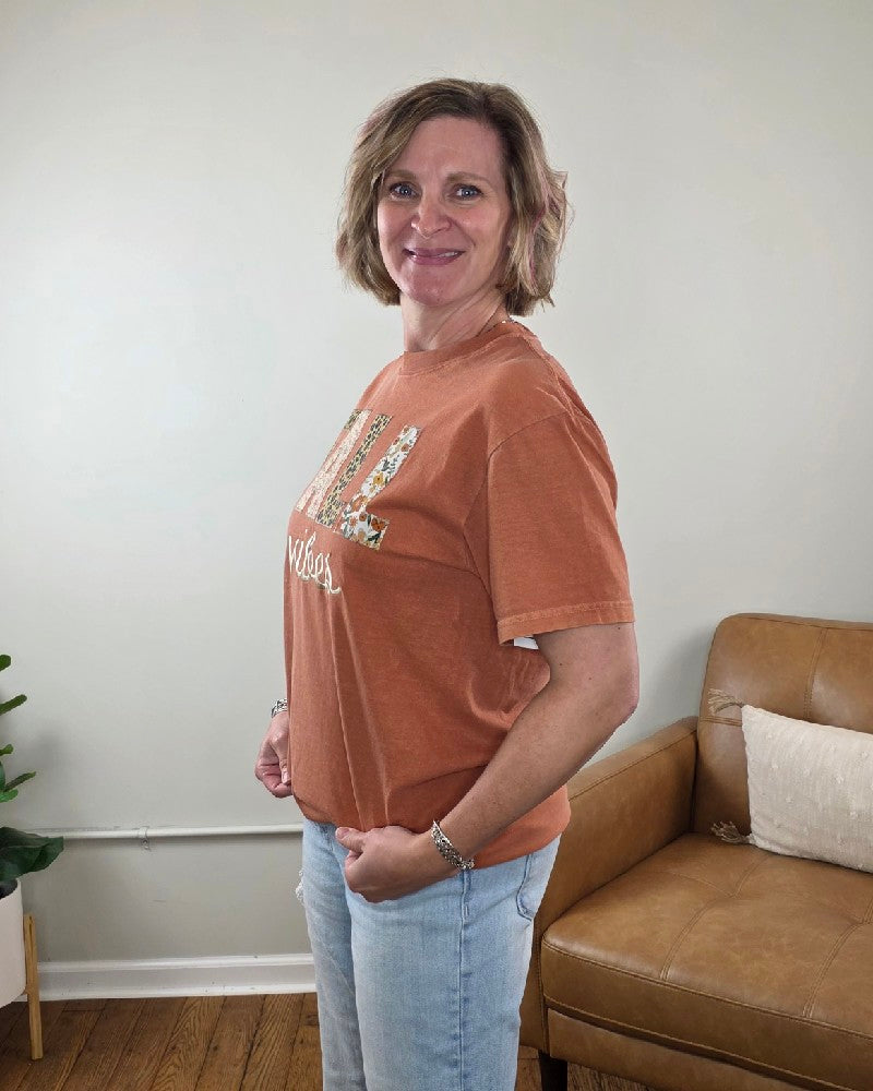 A woman wearing a rust-colored Fall Vibes Applique and Embroidered Tee featuring the word FALL in large, leopard floral applique block letters and the word vibes in cursive embroidery.