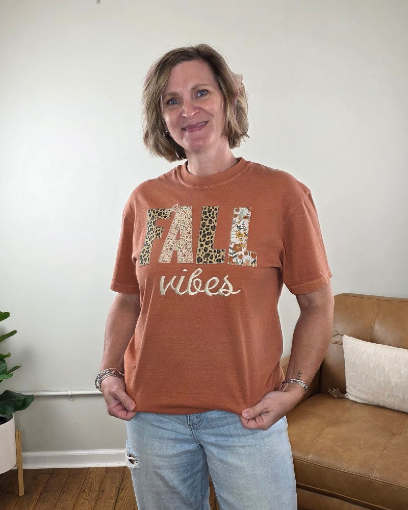 A woman wears a rust-colored Fall Vibes Applique and Embroidered Tee featuring FALL in leopard floral applique letters and vibes in white script.