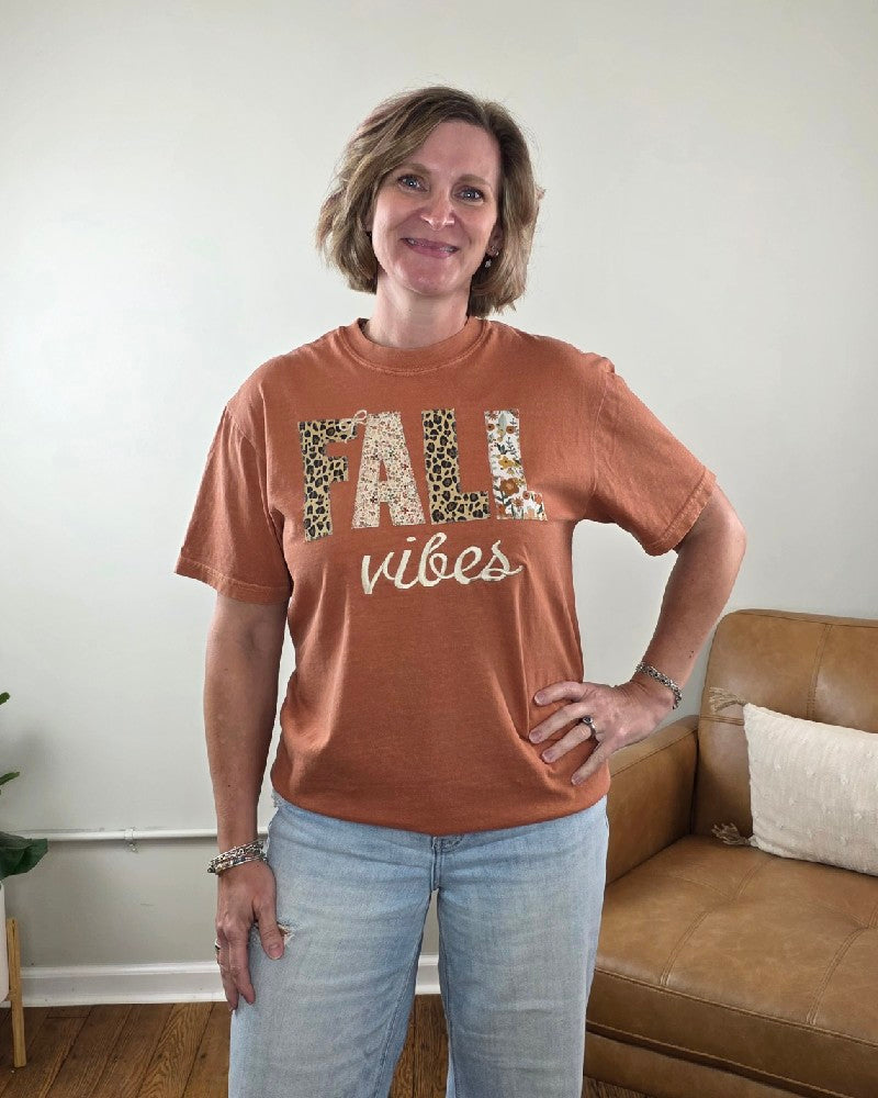 A woman wears a rust-colored Fall Vibes Applique and Embroidered Tee from Cosmos + Clover Boutique, featuring FALL in large patterned letters and vibes in white cursive script.