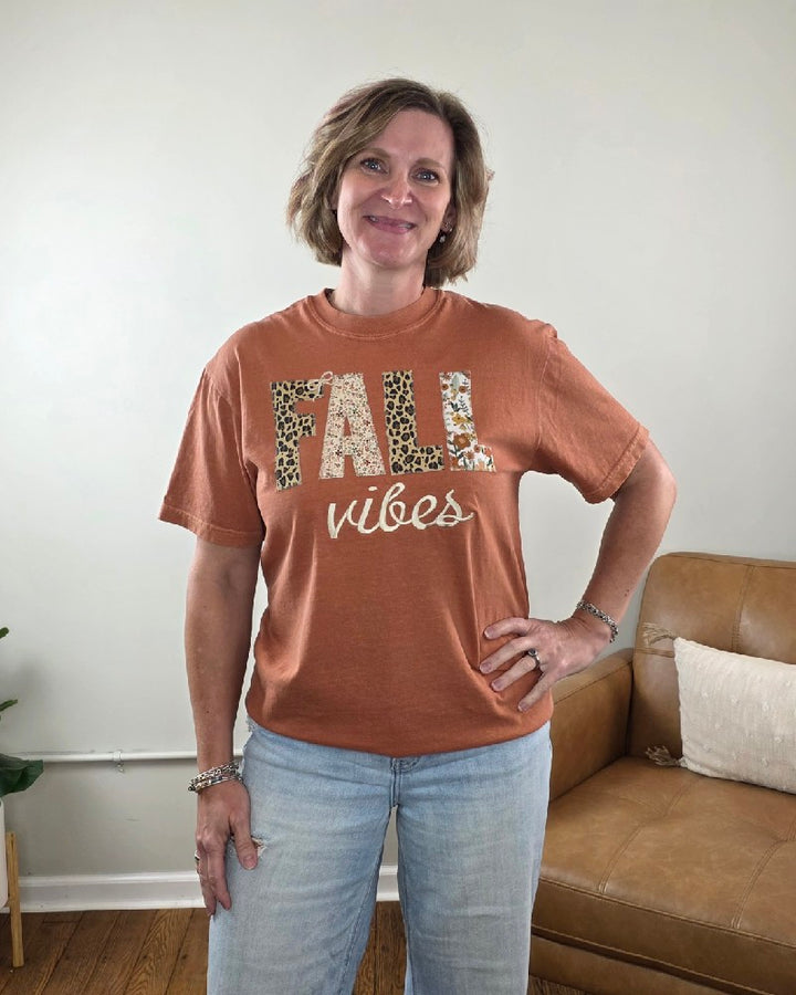 A woman wears a rust-colored Fall Vibes Applique and Embroidered Tee from Cosmos + Clover Boutique, featuring FALL in large patterned letters and vibes in white cursive script.