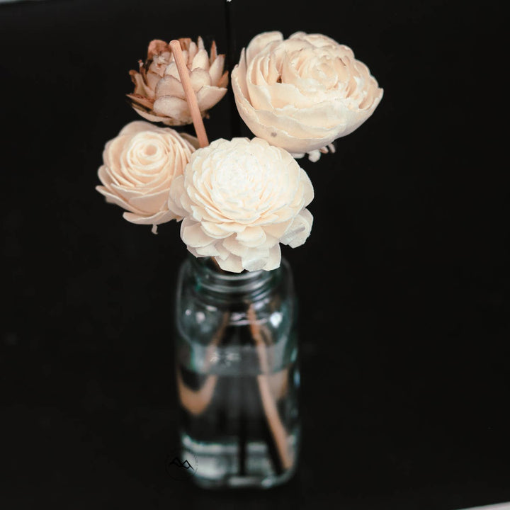 The Mahogany &amp; Teakwood - Flower Reed Diffuser by Cedar Mountain Candle features four pale cream-colored artificial flowers, natural rattan reeds, and water displayed in a clear glass bottle, set against a solid black background.