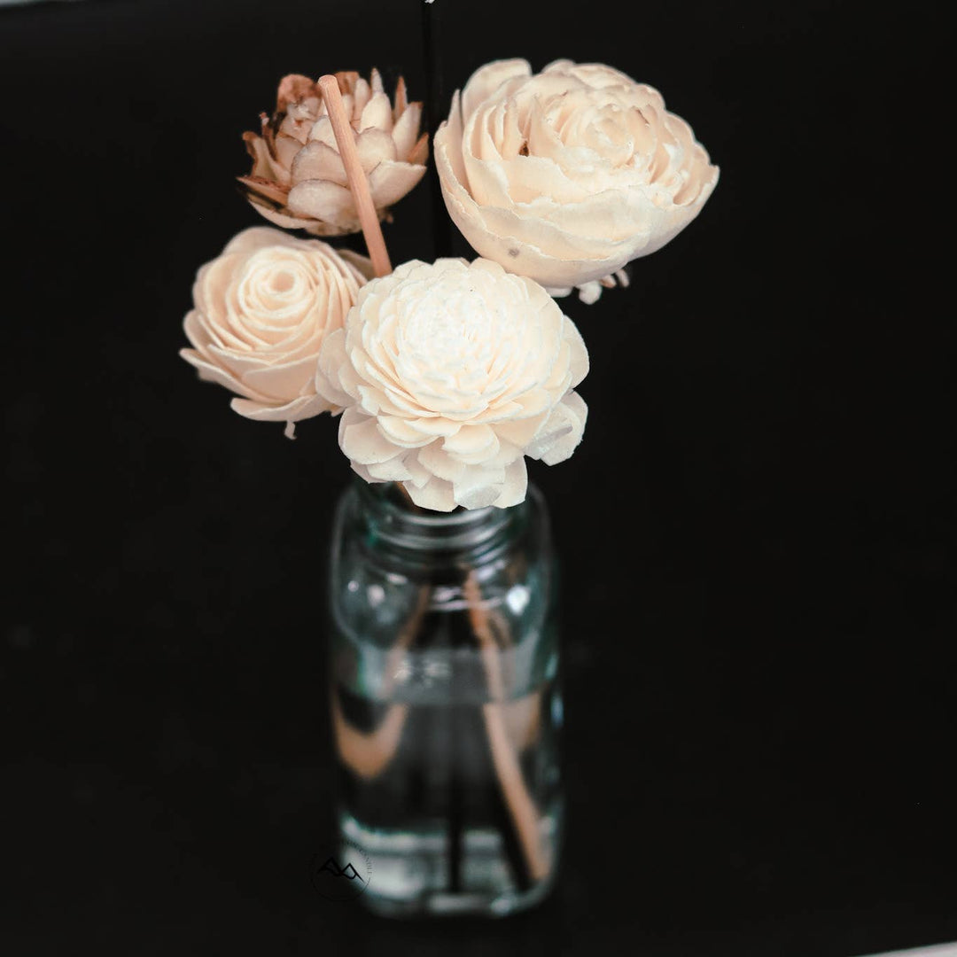 Four cream-colored sola wood flower reeds with layered petals from the Cedar Mountain Candle Cascading Waterfall Flower Reed Diffuser are arranged in a glass bottle filled with water, set against a solid black background.