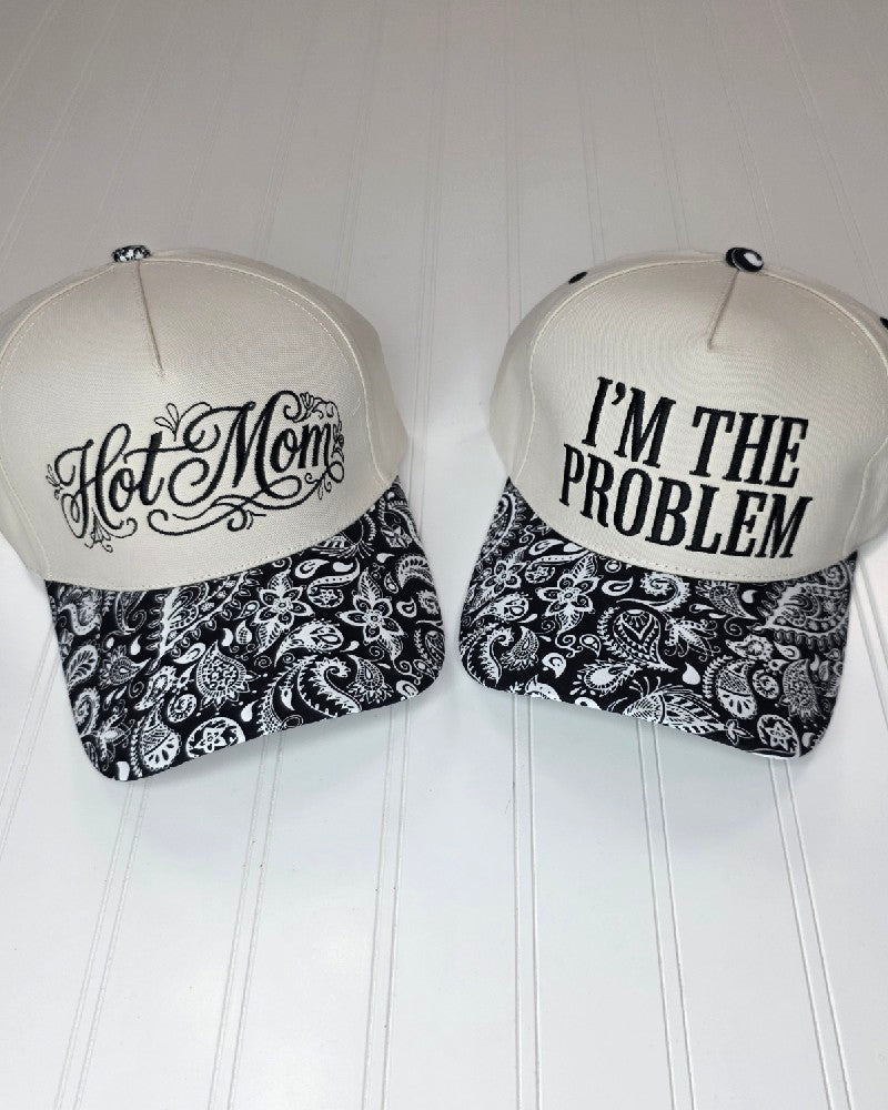 Two ZENANA Embroidered Hats in a classic white and black paisley design sit side by side on a white surface. One cap showcases "Hot Mom" in elegant script, while the other is adorned with "IM THE PROBLEM" in bold block letters—making these stylish hats from ZENANA the perfect pick for anyone who loves fun sayings and standout headwear.