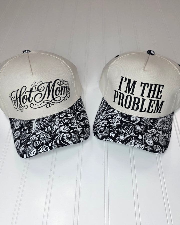 Two ZENANA Embroidered Hats in a classic white and black paisley design sit side by side on a white surface. One cap showcases "Hot Mom" in elegant script, while the other is adorned with "IM THE PROBLEM" in bold block letters—making these stylish hats from ZENANA the perfect pick for anyone who loves fun sayings and standout headwear.