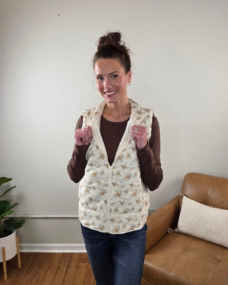 A woman wears a cream, floral-printed Staccato Isla Floral Printed Zip Up Vest over a brown long-sleeve shirt and jeans.