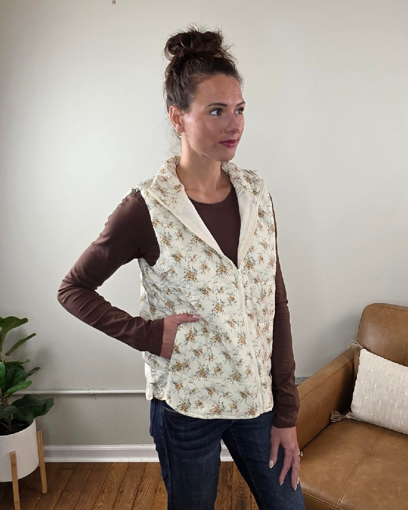 A woman wears a cream-colored floral printed Staccato Isla Floral Printed Zip Up Vest over a brown long-sleeved shirt and blue jeans.