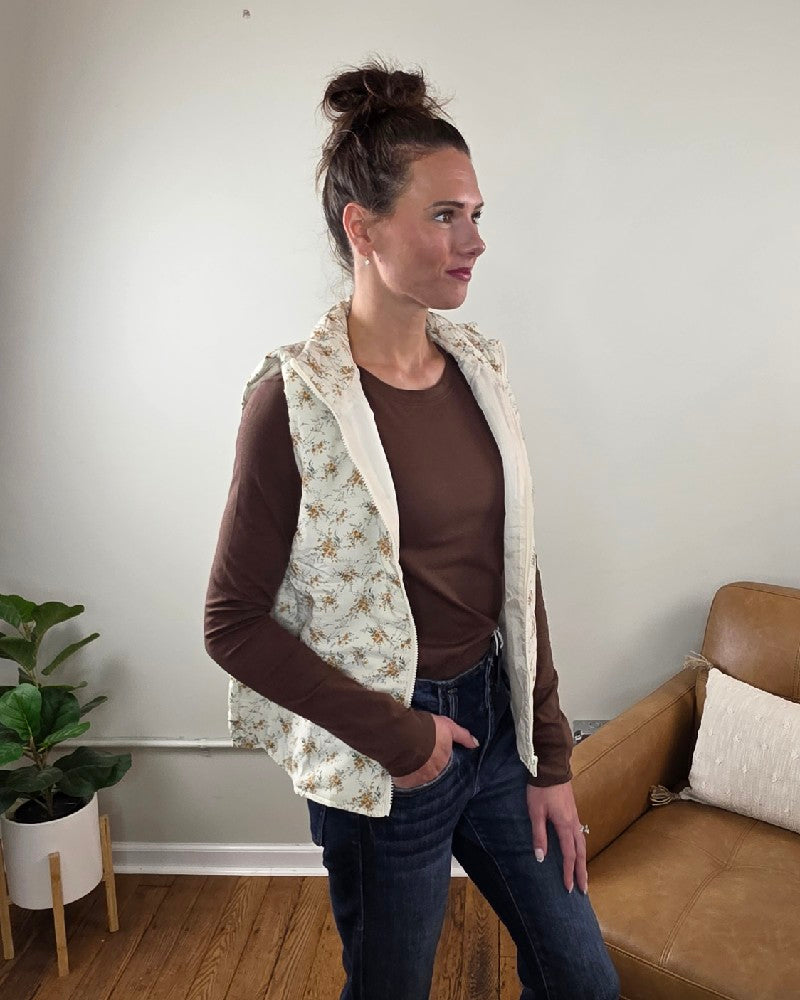 A woman wears a white floral printed Staccato Isla Floral Printed Zip Up Vest over a brown long-sleeved shirt and blue jeans.