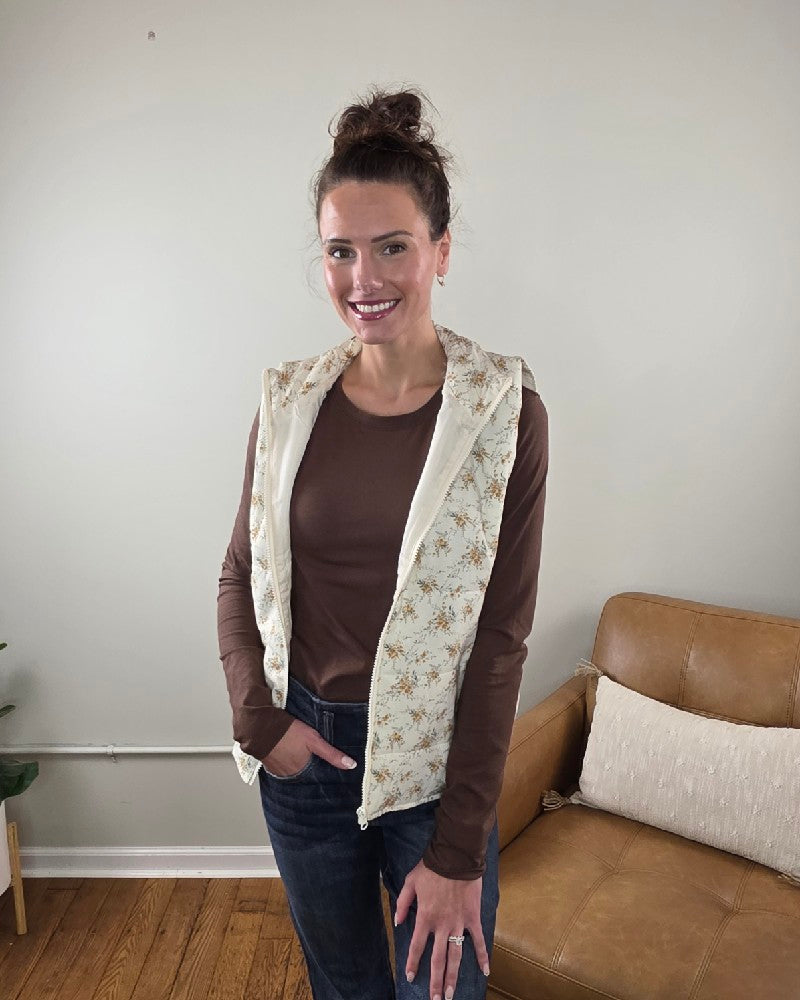 A woman wears a sleeveless, cream-colored floral printed Staccato Isla Floral Printed Zip Up Vest over a brown long-sleeved shirt and blue jeans.