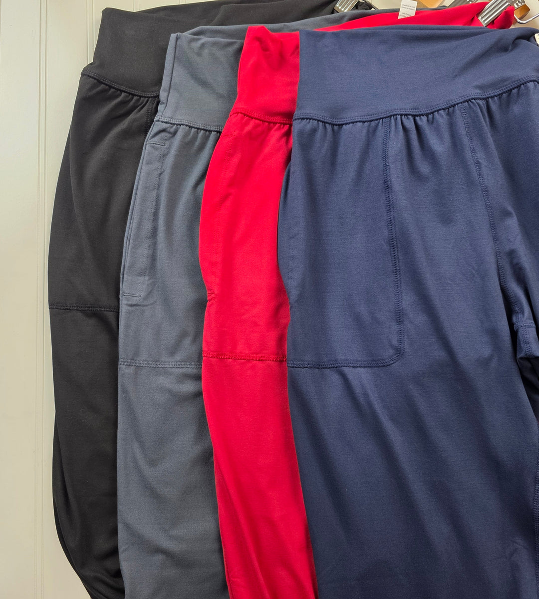 Four pairs of Rae Mode Cassie Joggers with gathered waistbands in black, charcoal gray, bright red, and navy blue hanging on a rack.