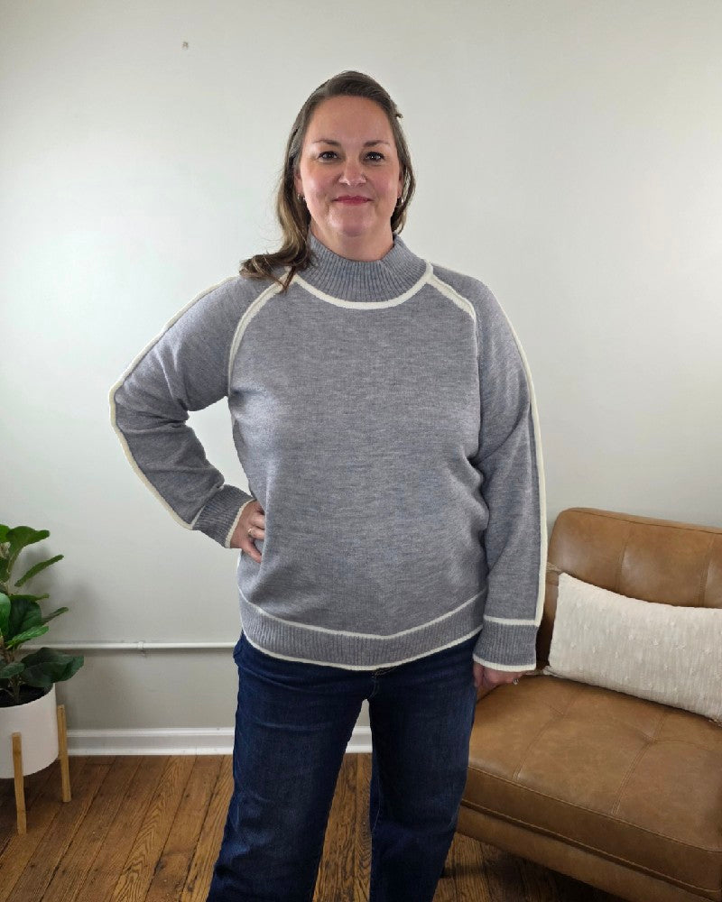 A woman wears a grey Kathy sweater with a mock neck and white trim detailing along the sleeves and hem.