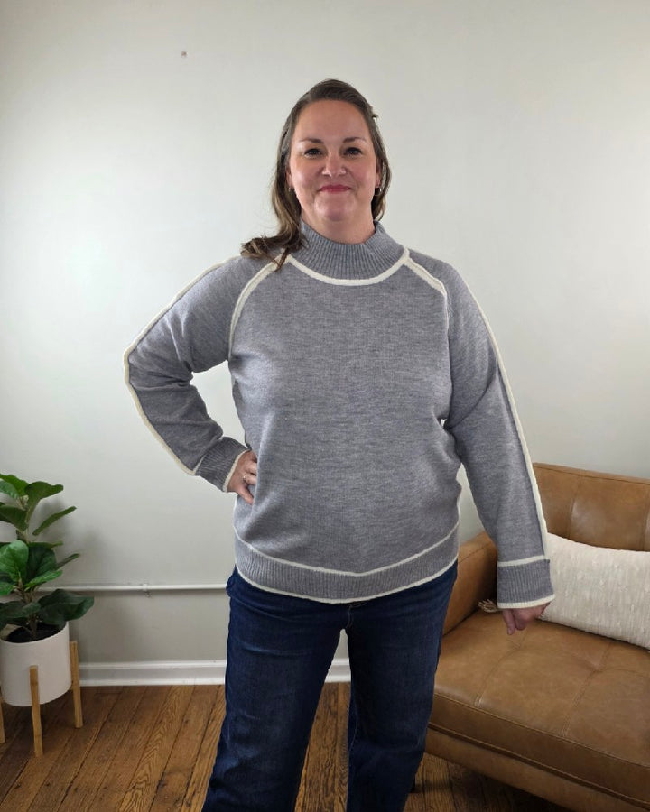 A woman wears a grey Kathy Sweater by Lime N Chili, Inc with a mock neck and contrasting white trim along the shoulders, cuffs, and hem, paired with blue jeans.