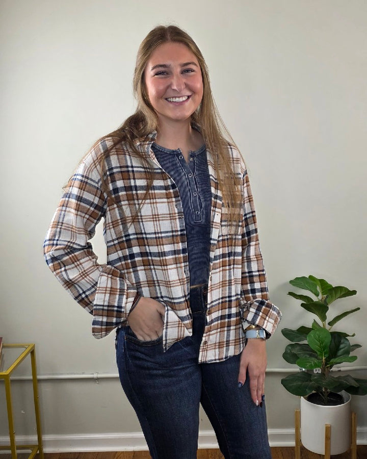 A smiling woman wearing a brown, white, and blue Kimberly Plaid Flannel Long Sleeve over a dark blue henley top and dark jeans.