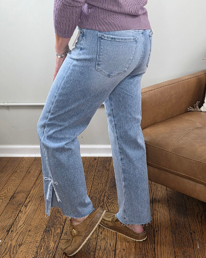A person wearing light blue Laura Crop Straight Jeans with Bow Embroidery from Risen, a lavender sweater, and tan moccasins stands on a wooden floor next to a brown leather couch.