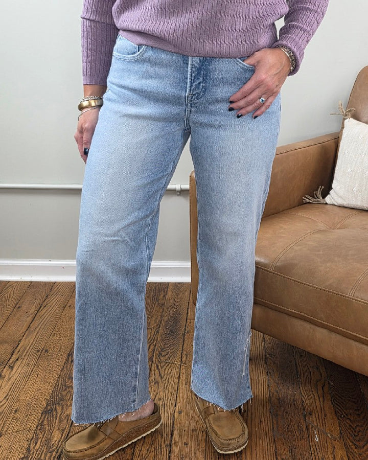 A woman wearing lavender knitwear and tan loafers stands next to a brown leather chair on a wooden floor, showcasing the Risen Laura Crop Straight Jeans with Bow Embroidery. Only her lower body and part of her torso are visible.