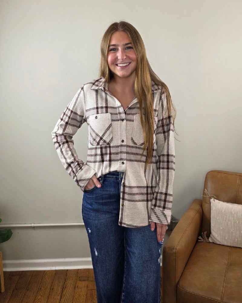 A woman with long hair smiles while standing indoors, wearing the Thread & Supply Lewis Plaid Button Down Flannel in cream and brown, paired with blue jeans. She stands near a tan armchair and a light-colored wall, with wood flooring visible.