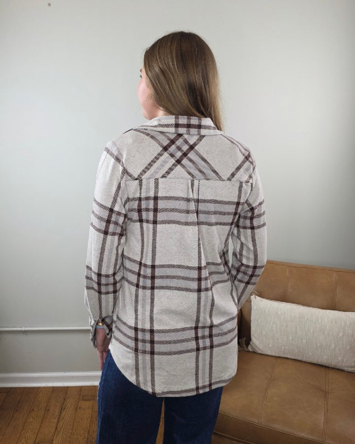 A person with long hair, wearing the Thread &amp; Supply Lewis Plaid Button Down Flannel in cream and brown and blue jeans, stands with their back to the camera in a simple room with a tan couch and wooden floor.