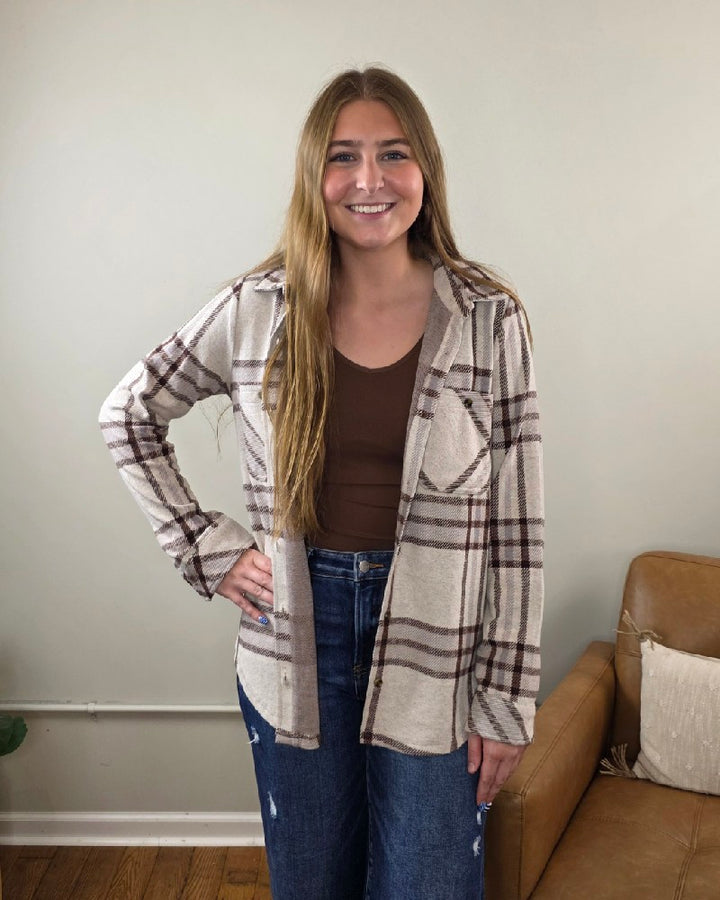 A woman with long light brown hair is smiling and standing indoors next to a tan couch against a plain wall. She wears a brown top, blue jeans, and the Thread &amp; Supply Lewis Plaid Button Down Flannel in cream and brown.