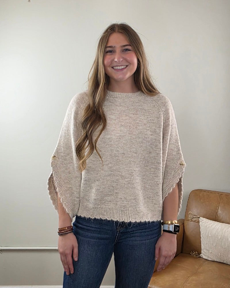 A woman with long brown hair, dressed in the LovLov Liora Soft Serenity Elegance Knit Pullover and jeans, smiles while standing indoors next to a tan couch against a light-colored wall.