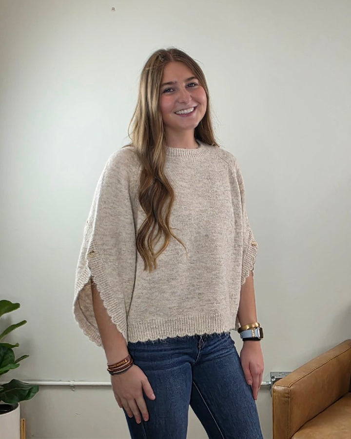 A woman with long wavy hair, dressed in the LovLov Liora Soft Serenity Elegance Knit Pullover—a relaxed silhouette sweater in loose beige knit—and blue jeans, stands smiling in a light-filled room with a plant and a tan chair visible.