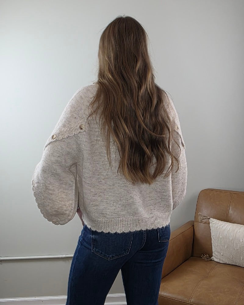 A woman with long wavy brown hair stands with her back to the camera, wearing the LovLov Liora Soft Serenity Elegance Knit Pullover and blue jeans in a room with a tan leather chair.