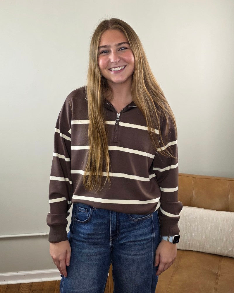 A woman with long brown hair smiles while standing indoors, wearing the Thread &amp; Supply Loma Striped Quarter Zip Pullover in a relaxed fit with brown and white stripes, paired with blue jeans. A beige couch and a light-colored wall are visible in the background.