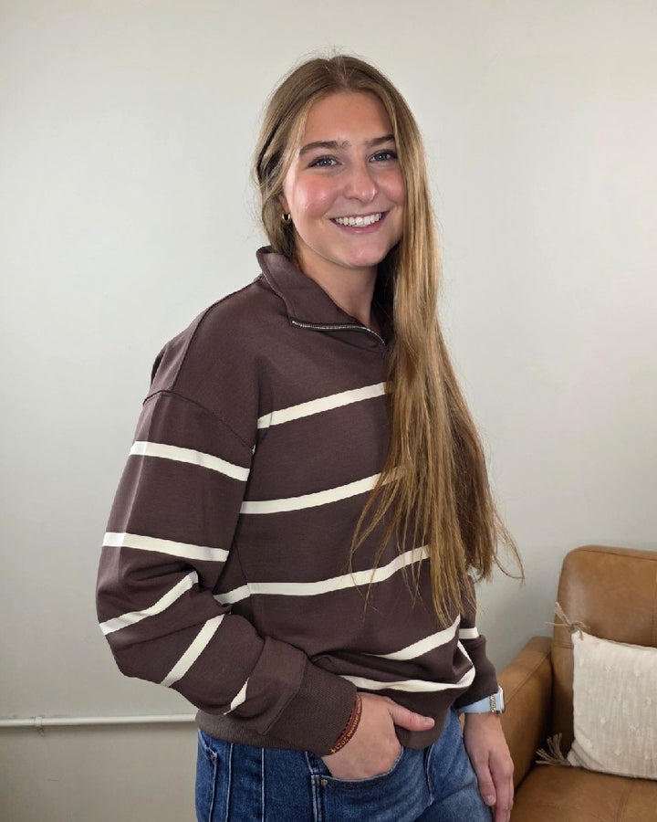A young woman with long, light brown hair wears the Thread &amp; Supply Loma Striped Quarter Zip Pullover and blue jeans, standing and smiling indoors near a tan couch with a white pillow—showcasing the ideal transitional layer for cozy comfort.