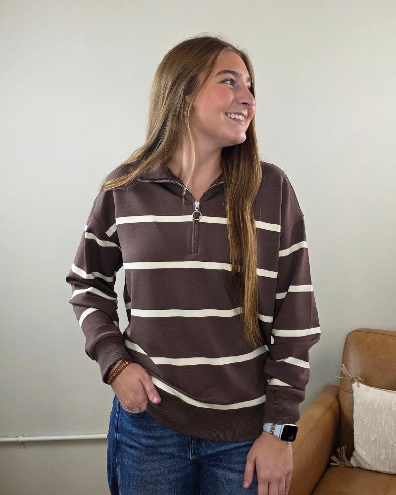 A woman with long brown hair is seen indoors by a tan couch, smiling and looking to her left, wearing the Thread & Supply Loma Striped Quarter Zip Pullover in a relaxed fit brown and white stripe, paired with blue jeans and a smartwatch. This versatile Loma pullover makes the perfect transitional layer for any casual outing.