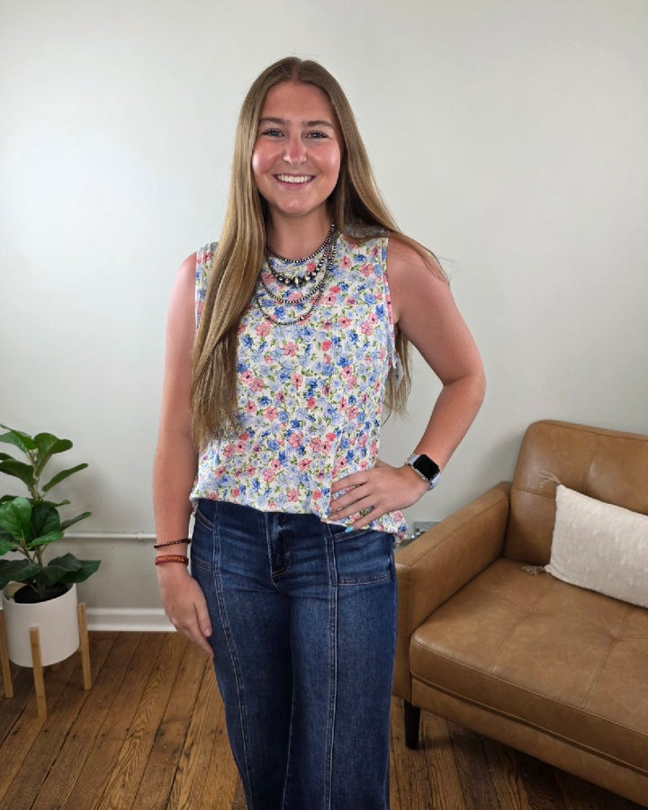 A woman with long brown hair smiles indoors, wearing a Haptics Maizy Floral Print Tank Top, blue jeans, a beaded necklace, and a smartwatch. Behind her are a tan leather chair, white pillow, and potted plant.