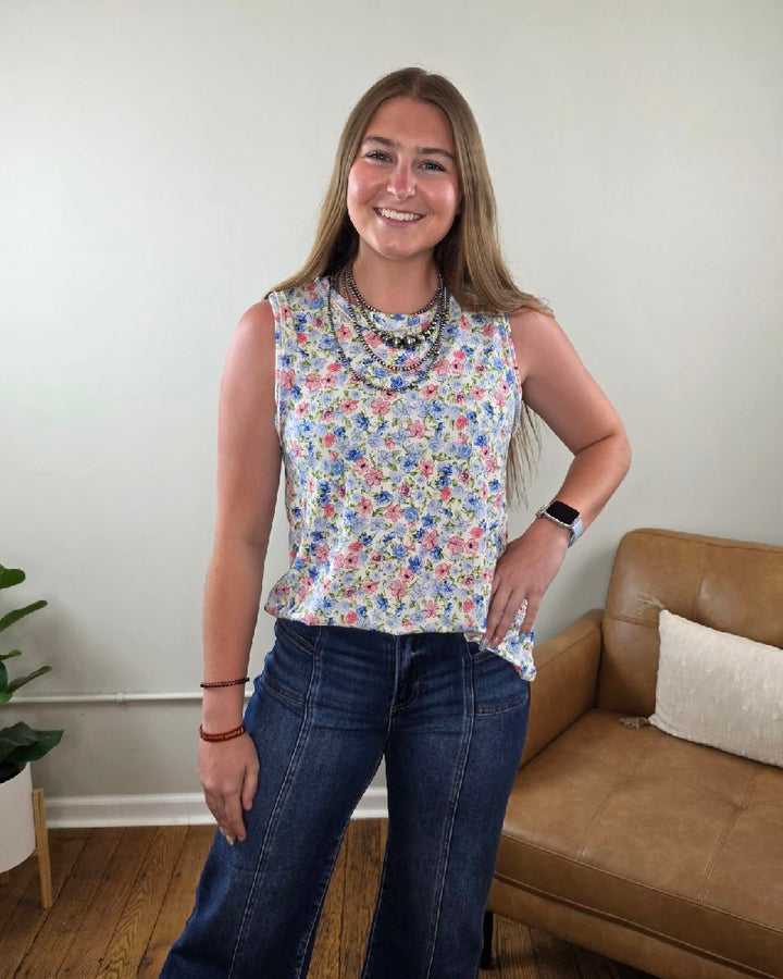 A woman with long brown hair smiles indoors, wearing a Haptics Maizy Floral Print Tank Top, layered necklaces, wide-leg jeans, and a smartwatch. Behind her is a brown couch with white pillows and a plant.