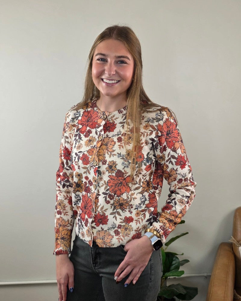 A woman wears the LovLov Megan Vintage Wildflower Cardigan, a cream-colored piece featuring a red, orange, and brown floral print.
