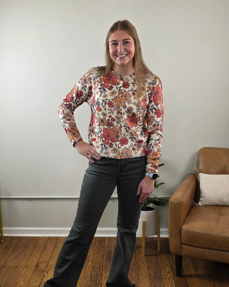 A person wears a cream LovLov Megan Vintage Wildflower Cardigan with red and orange floral patterns, buttoned up and paired with dark grey jeans.