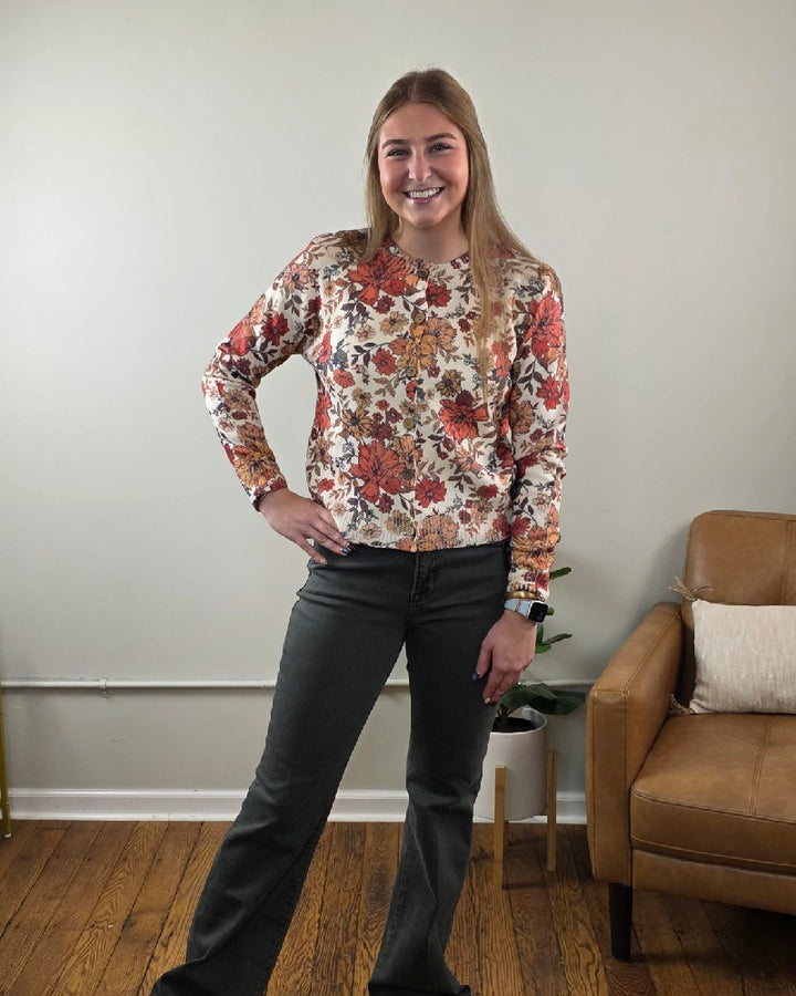 A person wears a cream LovLov Megan Vintage Wildflower Cardigan with red and orange floral patterns, buttoned up and paired with dark grey jeans.