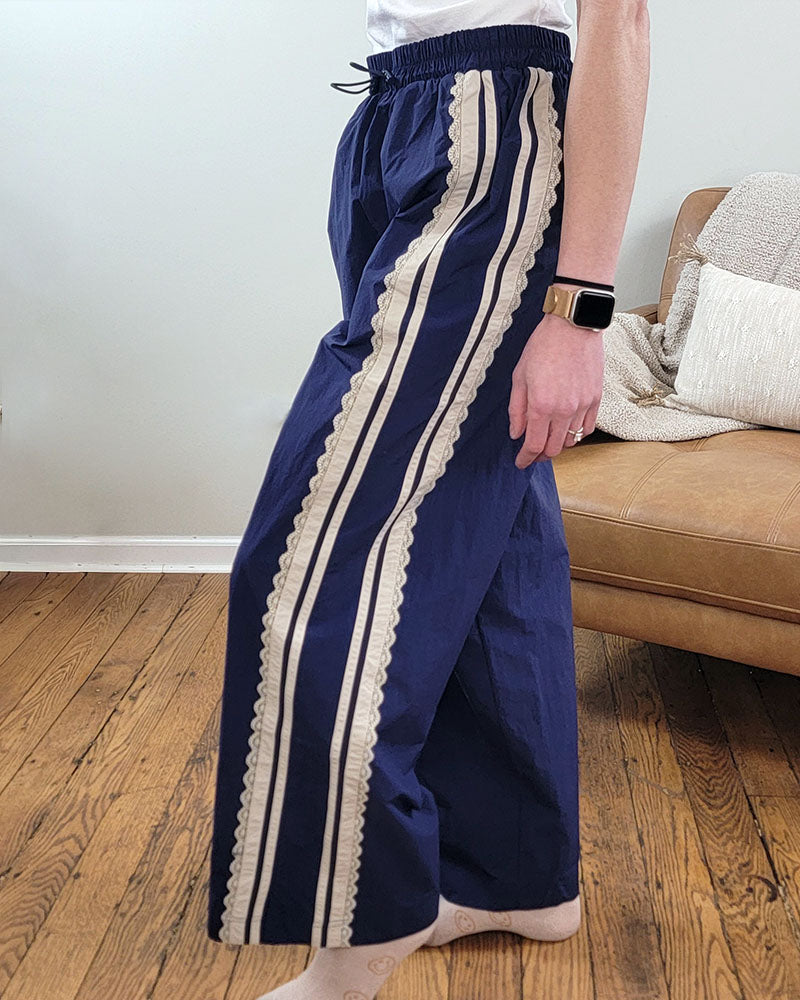 A person stands indoors on a wooden floor wearing Veveret’s Scarlett Striped Lace Pants—navy with beige side stripes and an elastic waistband—with a white top and smartwatch. A tan cushioned sofa is in the background.