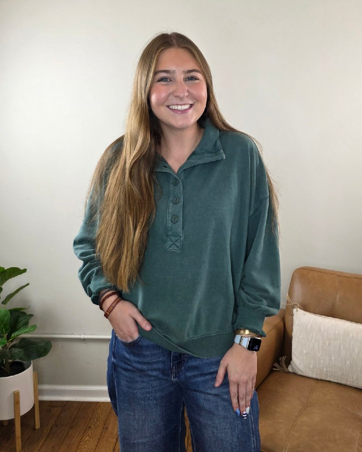 A smiling woman wearing a teal, long-sleeved Nora Button Front Sweatshirt and blue jeans.