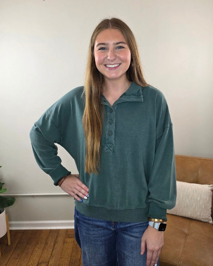 A woman wearing the dark green, button-down Nora Button Front Sweatshirt by HYFVE, Inc, featuring a relaxed fit, long sleeves, and a ribbed hem.