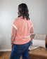 A person with wavy brown hair, wearing the ZENANA Brealyn Stripe Tee in pink and white and blue jeans, stands with their back to the camera beside a tan couch featuring a blanket and pillow.